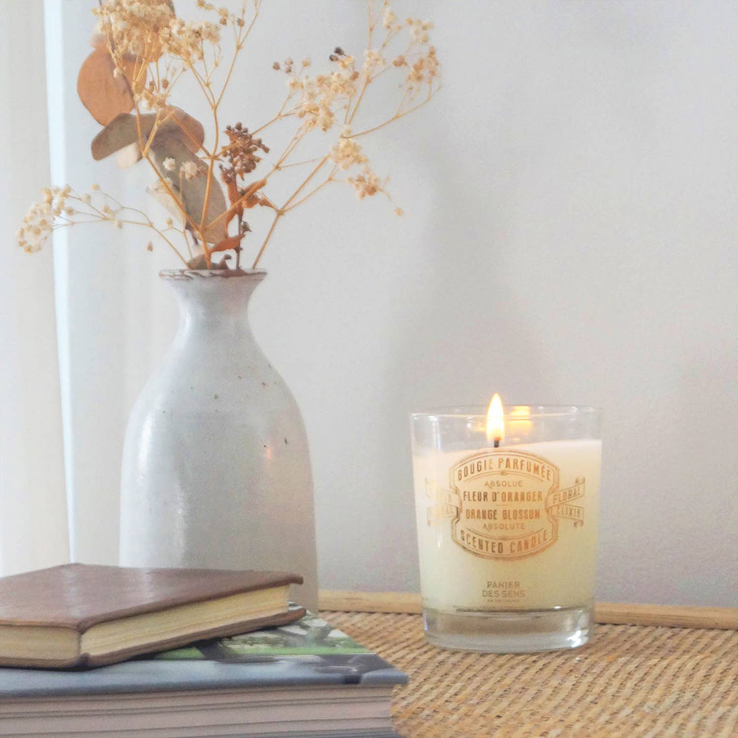 Die handgefertigte "Duftkerze | Orangenblüte 180 g" von Panier Des Sens brennt im Glas neben einer Keramikvase mit Trockenblumen und einem Notizbuch auf geflochtenem Untergrund, vorn liegt ein Bücherstapel.
