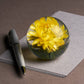 Ein silberner Stift und der Hafod Grange Briefbeschwerer mit Gelber Helichrysum ruhen auf grauem Papier auf einer braunen Oberfläche - das ideale Geschenk für Blumenliebhaber.