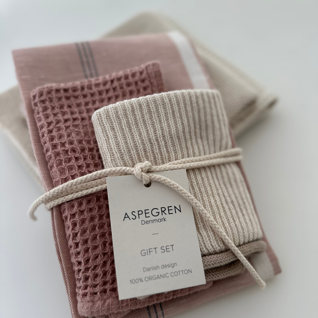 Ein Set aus drei Küchentüchern in warmen Rosatönen von Aspegren Denmark, hergestellt aus 100 % Bio-Baumwolle und als Geschenkset mit einer Schnur sorgfältig gefaltet, wird auf einer hellen Unterlage präsentiert.