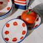 Eine frische Tomate liegt in einer kleinen Knoblauchreibe „Pomodoro“ von Alfar Tierra Cocida, die mit roten Tomatenmotiven verziert und im traditionellen Keramikstil handgefertigt ist. Daneben liegt ein weiteres passendes Stück von Alfar Tierra Cocida – eine handgefertigte Keramik-Reibplatte aus Spanien mit einer wunderschön strukturierten Oberfläche –, alles präsentiert auf einer weißen Tischdecke, die in helles Sonnenlicht getaucht ist.
