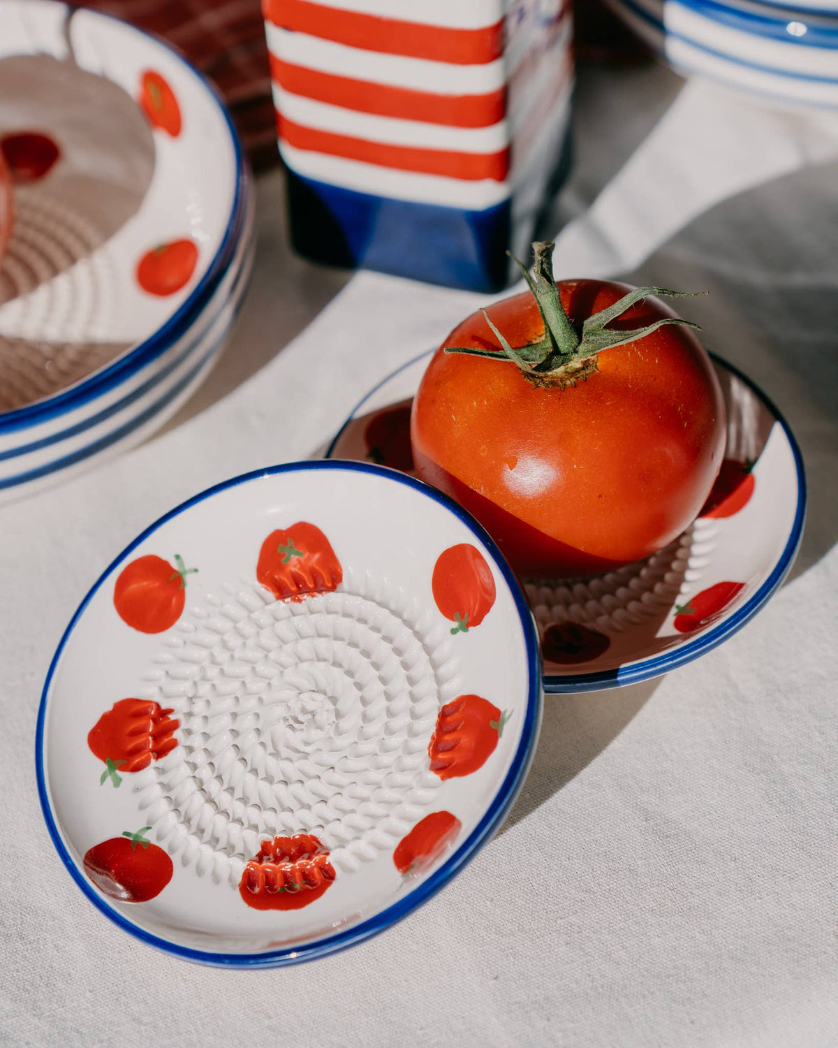 Eine frische Tomate liegt in einer kleinen Knoblauchreibe „Pomodoro“ von Alfar Tierra Cocida, die mit roten Tomatenmotiven verziert und im traditionellen Keramikstil handgefertigt ist. Daneben liegt ein weiteres passendes Stück von Alfar Tierra Cocida – eine handgefertigte Keramik-Reibplatte aus Spanien mit einer wunderschön strukturierten Oberfläche –, alles präsentiert auf einer weißen Tischdecke, die in helles Sonnenlicht getaucht ist.