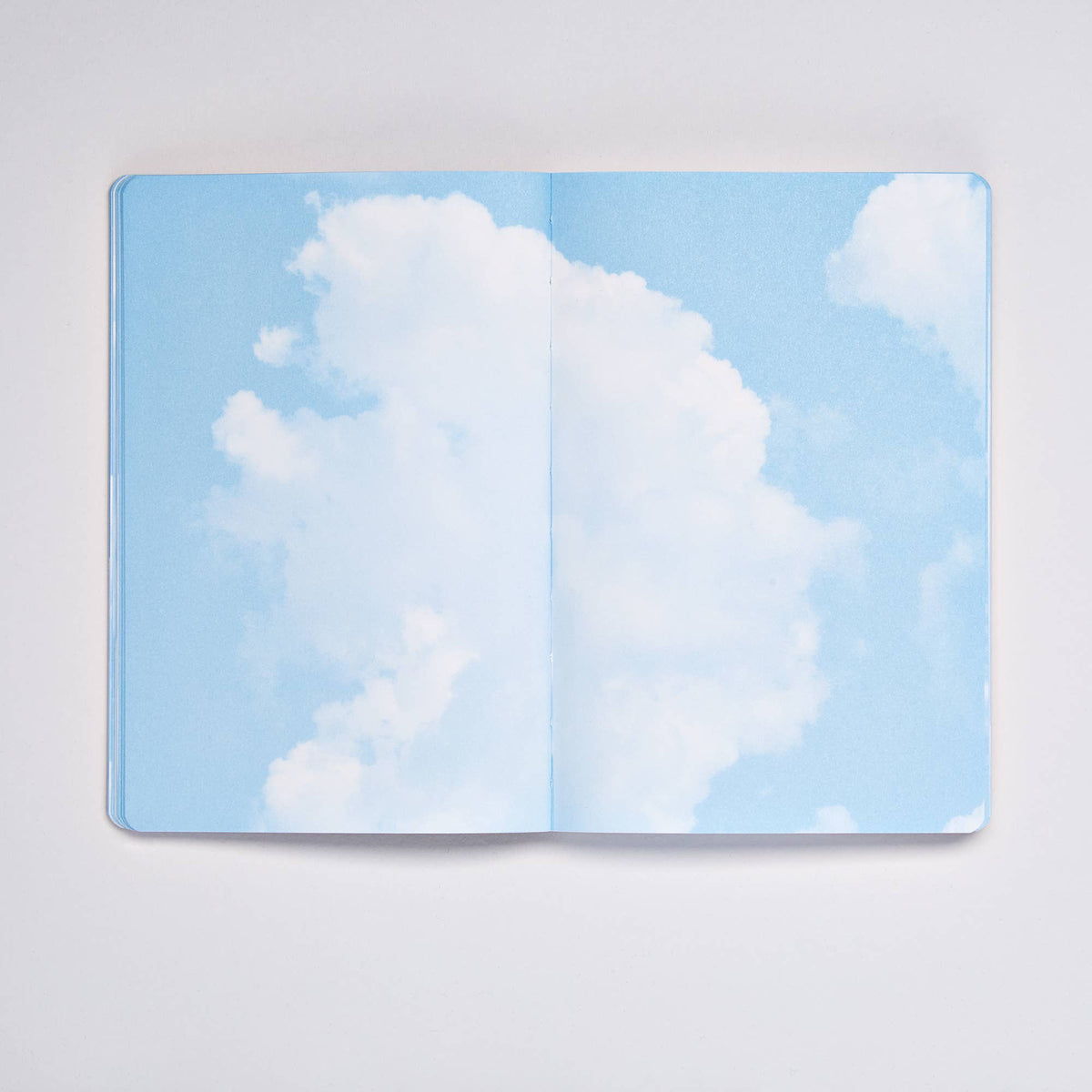 Ein offenes nuuna | „Cloud Blue“-Inspirationsbuch in Größe M mit 176 farbigen Seiten liegt auf einer weißen Fläche, wobei beide Seiten ein Bild des blauen Himmels und weißer Wolken der Marke nuuna zeigen – wodurch der Eindruck entsteht, man blicke in den Himmel.
