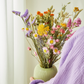 Eine Person in einem lavendelfarbenen Gewand hält vor einem hellen Hintergrund eine grüne Vase in der Hand, die mit den bunten Trockenblumen „Feldblumenstrauß“ (Größe S–M) aus der Kollektion „Wildflowers“ von Floriette gefüllt ist, die gelbe, rosa, orangefarbene und violette Blüten aufweisen.