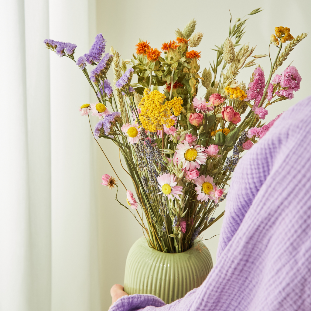 Eine Person in einem lavendelfarbenen Gewand hält vor einem hellen Hintergrund eine grüne Vase in der Hand, die mit den bunten Trockenblumen „Feldblumenstrauß“ (Größe S–M) aus der Kollektion „Wildflowers“ von Floriette gefüllt ist, die gelbe, rosa, orangefarbene und violette Blüten aufweisen.