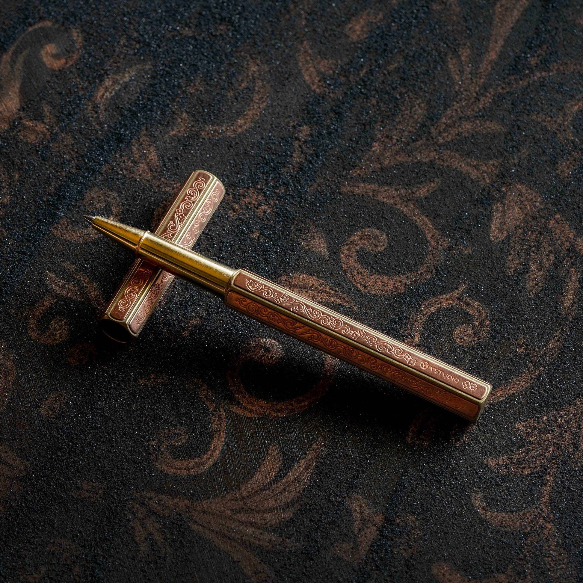 Der ENTREPÔT YSTUDIO Klassische Renaissance KAZARI KANAGU ORCHID Tintenroller mit filigraner Metallgravurkunst ruht auf einer dunkel gemusterten Oberfläche, wobei der unverschlossene Schaft und die Kappe in einem Kreuz angeordnet sind, um raffinierte Handwerkskunst zu präsentieren.