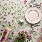 Eine Tischdecke mit Blumenmuster – wie beispielsweise die atemberaubende Tischdecke „Flora Danica Atlas“ von Koustrupco (350 x 145 cm) – bildet den Rahmen für die Tischdekoration, bestehend aus einem weißen Teller, einer Teetasse, Besteck, einem Glastablett mit einem aufgerollten Dessert, das mit Blumen garniert ist, sowie einem kleinen Strauß aus violetten Blumen und Grünpflanzen.