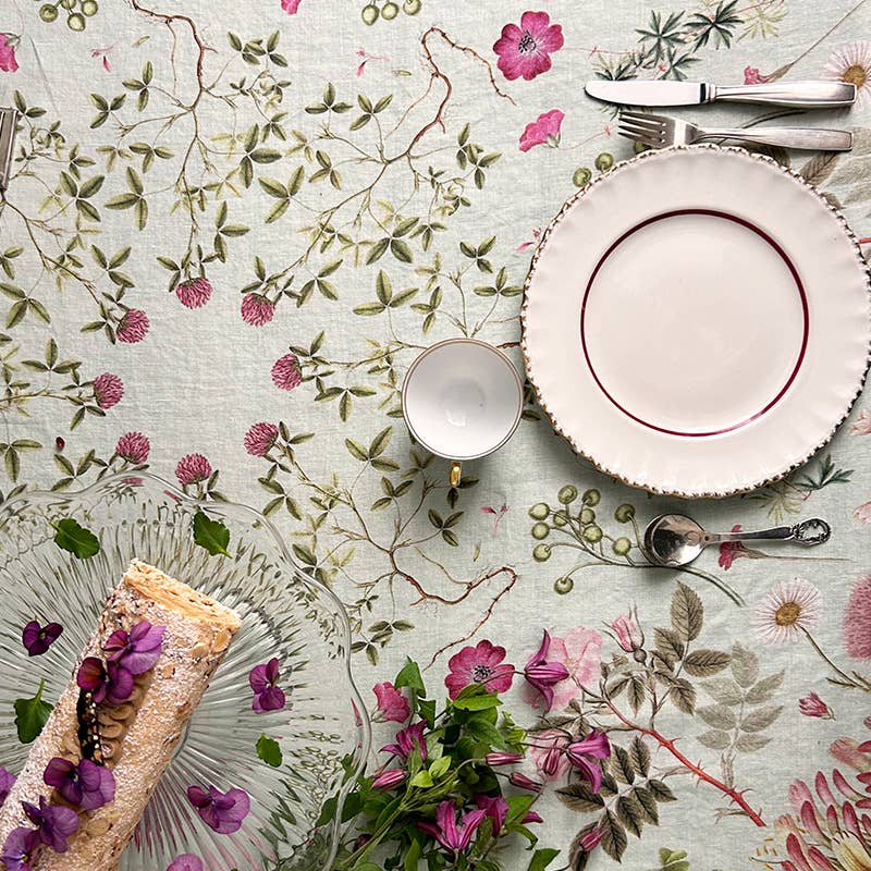 Eine Tischdecke mit Blumenmuster – wie beispielsweise die atemberaubende Tischdecke „Flora Danica Atlas“ von Koustrupco (350 x 145 cm) – bildet den Rahmen für die Tischdekoration, bestehend aus einem weißen Teller, einer Teetasse, Besteck, einem Glastablett mit einem aufgerollten Dessert, das mit Blumen garniert ist, sowie einem kleinen Strauß aus violetten Blumen und Grünpflanzen.