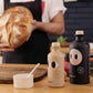 In einer Küche hält eine Person in einer Schürze Brot, während zwei stilvolle Flaschen von yiayia and friends - eine mit der Aufschrift "Natives Olivenöl Extra mit Knoblauch 200 ml" - neben einer kleinen Schüssel mit Löffel stehen, die die Essenz der mediterranen Küche einfangen.