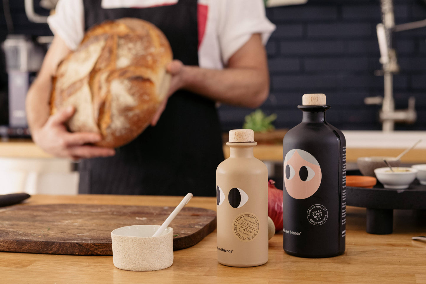 In einer Küche hält eine Person in einer Schürze Brot, während zwei stilvolle Flaschen von yiayia and friends - eine mit der Aufschrift "Natives Olivenöl Extra mit Knoblauch 200 ml" - neben einer kleinen Schüssel mit Löffel stehen, die die Essenz der mediterranen Küche einfangen.