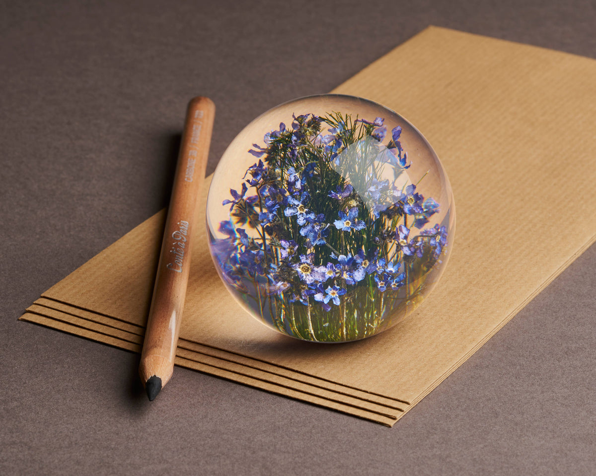Ein Holzstift und der „Vergissmeinnicht“-Briefbeschwerer von Hafod Grange Ltd – eine Kugel aus klarem Glas, gefüllt mit blauen Frühlingsblumen (Myosotis sylvatica) – liegen auf einem Stapel brauner Papierbögen, der auf einer dunklen Unterlage ruht.