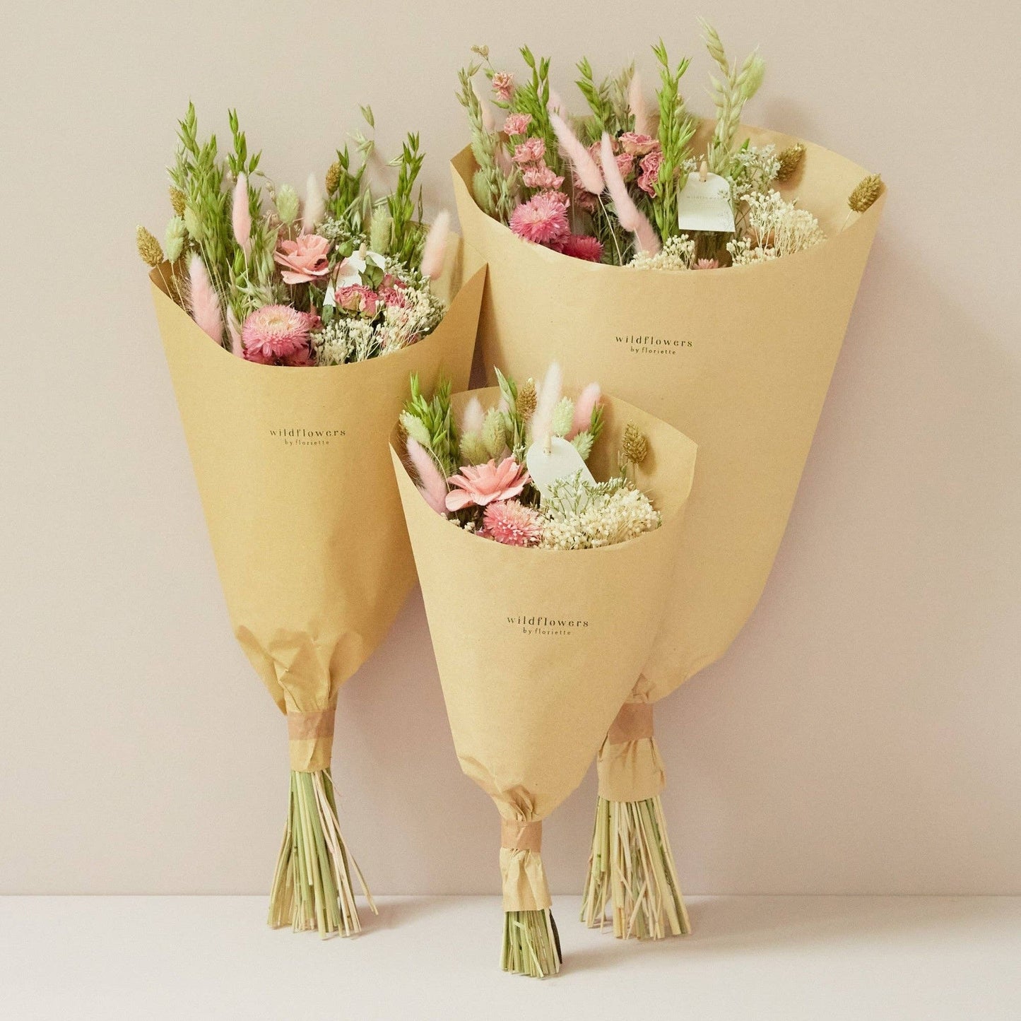 Drei in braunes Papier eingewickelte Sträuße aus rosa und weißen Trockenblumen mit grünem Laub sind vor einem hellbeigen Hintergrund arrangiert. Jeder Strauß trägt die Aufschrift „Wildflowers“ und verkörpert den Charme des Wiesenstraußes, der jeder Umgebung die „Rosa Eleganz“ der Wildflowers by Floriette | Trockenblumen Feldstrauß in Größe S verleiht.