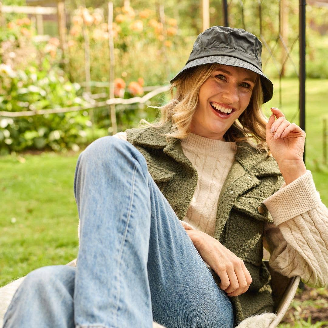 Eine lächelnde Frau, die den schwarzen Krempenhut „Wachstropfen“ von Tweedmill Textiles trägt und dazu einen cremefarbenen Pullover, eine grüne Weste und Jeans, sitzt draußen auf dem Rasen und wirkt entspannt und fröhlich.