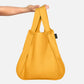 Eine Hand hält die Notabag | „Original Golden“, eine vielseitige Notabag-Kombination aus Tasche und Rucksack mit breiten Tragegriffen, vor einem schlichten weißen Hintergrund.