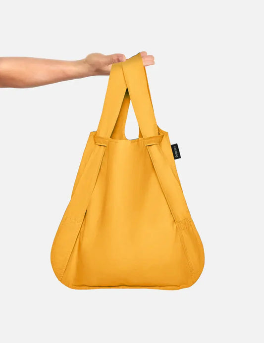 Eine Hand hält die Notabag | „Original Golden“, eine vielseitige Notabag-Kombination aus Tasche und Rucksack mit breiten Tragegriffen, vor einem schlichten weißen Hintergrund.
