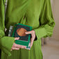 Eine Person in einem leuchtend grünen Kleid hält mehrere Tinne Mia Notizbücher mit Knopf 'Leinen Smaragd', wobei die bunten Einbände und ihre Hände hervorgehoben werden; ein Gesicht ist nicht zu sehen.