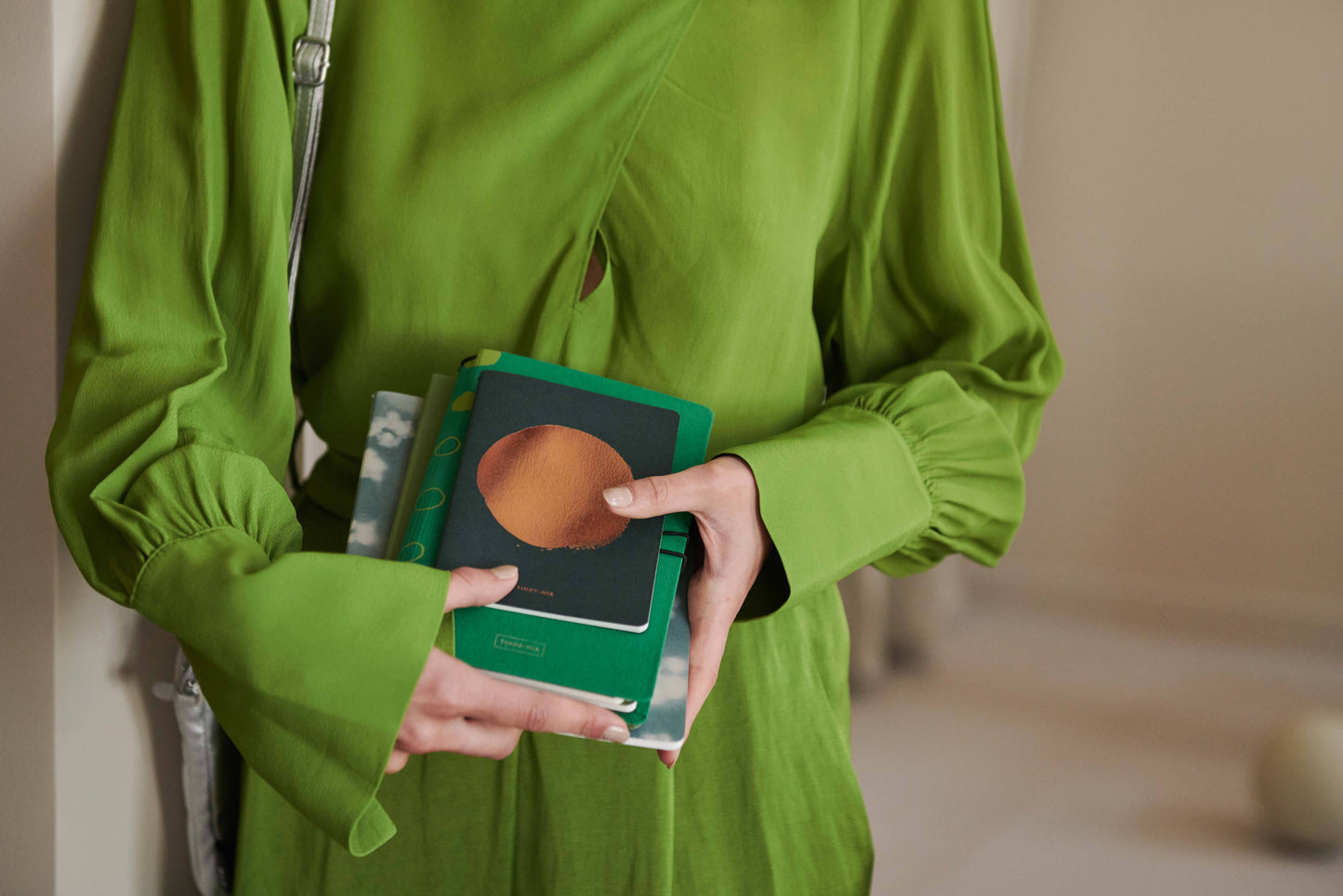 Eine Person in einem leuchtend grünen Kleid hält mehrere Tinne Mia Notizbücher mit Knopf 'Leinen Smaragd', wobei die bunten Einbände und ihre Hände hervorgehoben werden; ein Gesicht ist nicht zu sehen.