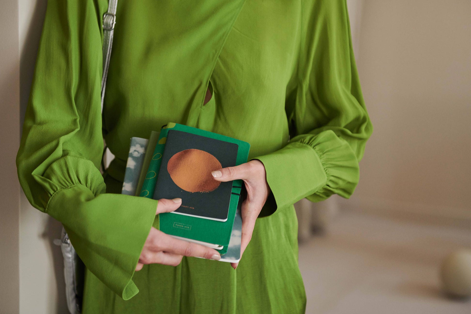 Eine Person in einem leuchtend grünen Kleid hält mehrere Tinne Mia Notizbücher mit Knopf 'Leinen Smaragd', wobei die bunten Einbände und ihre Hände hervorgehoben werden; ein Gesicht ist nicht zu sehen.