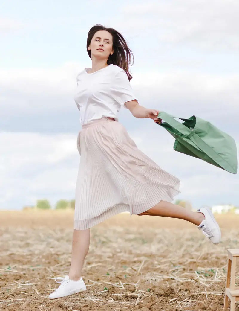 Eine Frau in einem weißen Hemd, einem Faltenrock und weißen Sneakers springt auf einem trockenen Feld und hält dabei ihre Notabag | Rucksack und Tasche in einem „Olive“ von Notabag in der Hand – eine innovative, wiederverwendbare Rucksack-Tragetasche. Der Himmel ist teilweise bewölkt, und sie blickt selbstbewusst nach vorne.