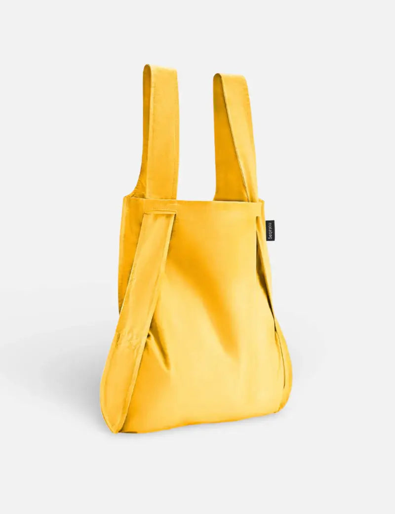 Die „Original Golden“ von Notabag ist eine leuchtend gelbe, umweltfreundliche Kombination aus Tragetasche und Rucksack mit breiten Tragegriffen und einem kleinen schwarzen Etikett, die aufrecht vor einem schlichten weißen Hintergrund abgebildet ist.