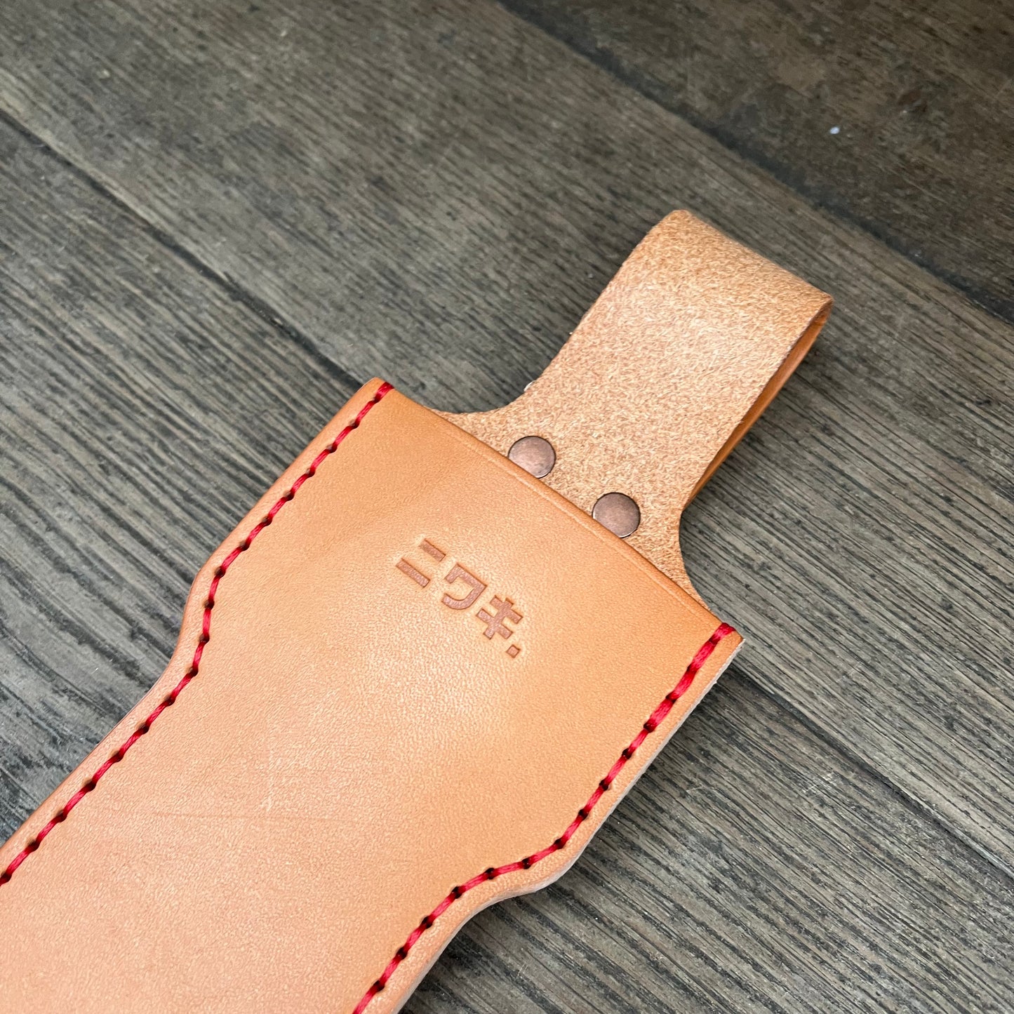 Eine hellbraune Niwaki | 'Long Holster' Lederschutzhülle für große Scheren mit roten Nähten und zwei Metallnieten liegt auf einer hölzernen Oberfläche, auf der ein japanischer Text für den stilvollen Schutz großer Werkzeuge eingeprägt ist.
