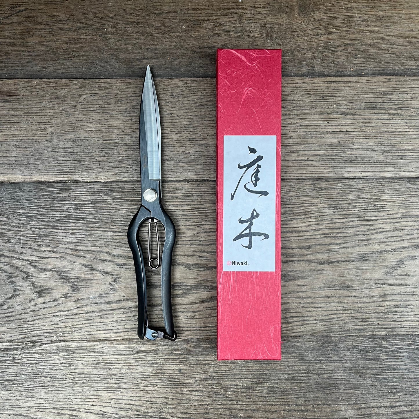 Ein Paar Niwaki 'Sentei Topiary Clippers' Buchsbaumschere mit Schlaufengriffen ruht neben einer hohen roten Schachtel mit japanischer Kalligrafie, beide von Niwaki, ausgestellt auf einer Holzfläche.