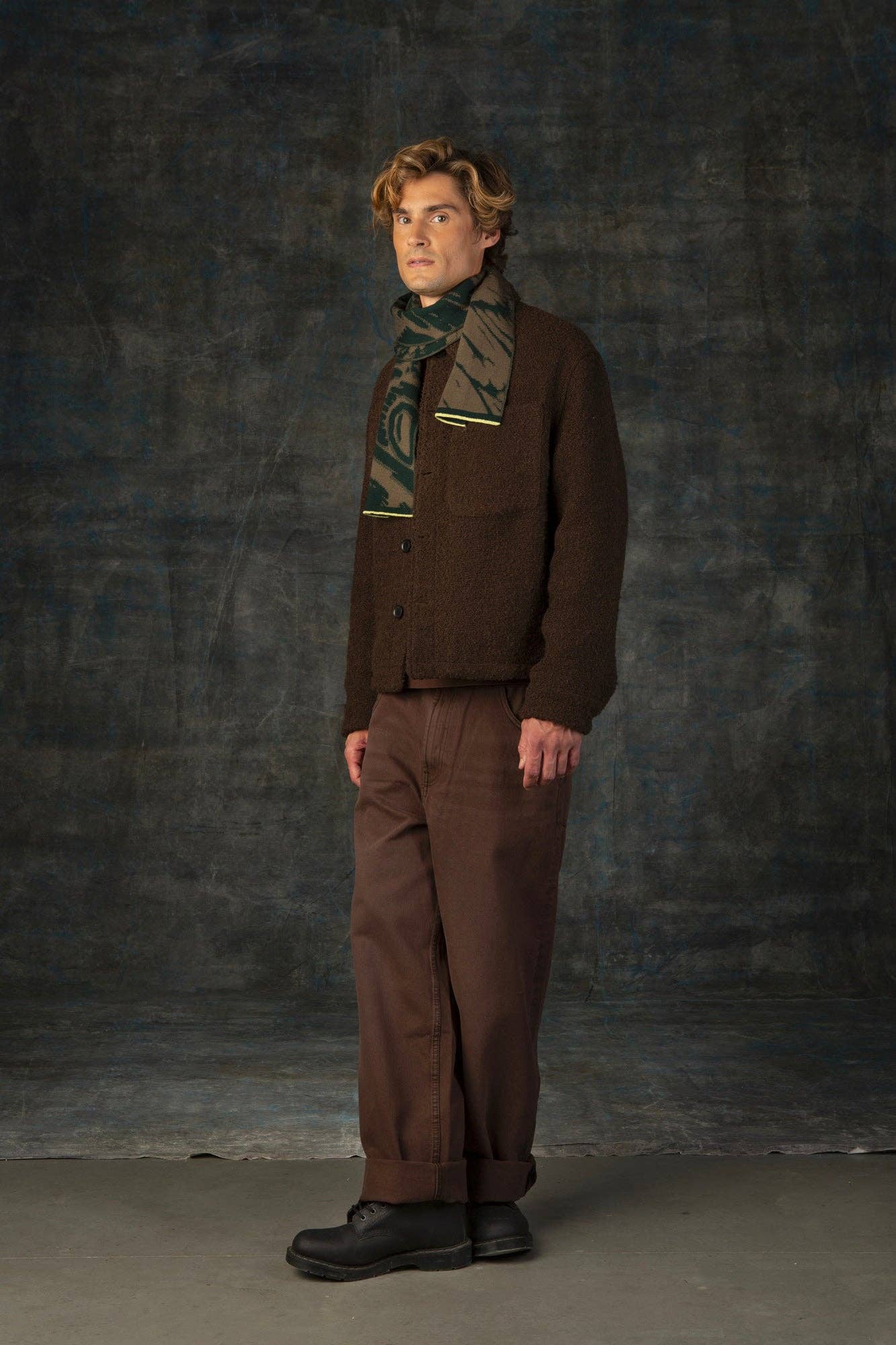Ein Mann steht vor einem dunkel strukturierten Hintergrund und trägt eine braune Strickjacke, eine braune Hose, schwarze Schuhe und den Furcula Wald Mini Schal aus Merinowolle von Wolvis. Er blickt mit neutraler Miene nach vorne.