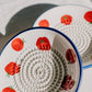 Zwei Teller der Serie „Alfar Tierra Cocida | Kleine Keramik Knoblauchreibe ‚Pomodoro‘“, handgefertigt in Spanien mit blauen Rändern und Tomatenmotiven sowie strukturierten spiralförmigen Mittelpartien, sind auf einer weißen Fläche ausgestellt, auf der das Sonnenlicht sanfte Schatten wirft.
