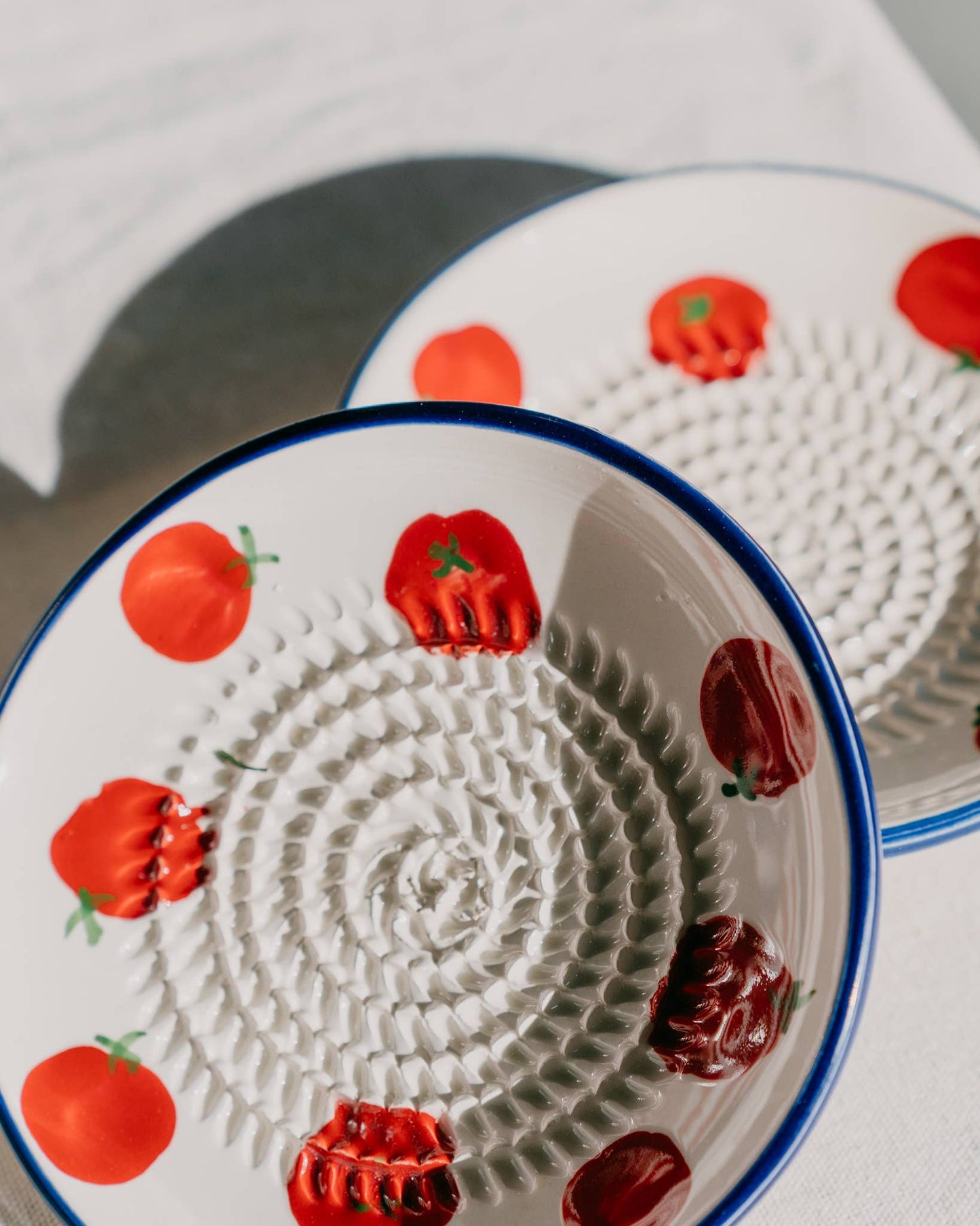 Zwei Teller der Serie „Alfar Tierra Cocida | Kleine Keramik Knoblauchreibe ‚Pomodoro‘“, handgefertigt in Spanien mit blauen Rändern und Tomatenmotiven sowie strukturierten spiralförmigen Mittelpartien, sind auf einer weißen Fläche ausgestellt, auf der das Sonnenlicht sanfte Schatten wirft.