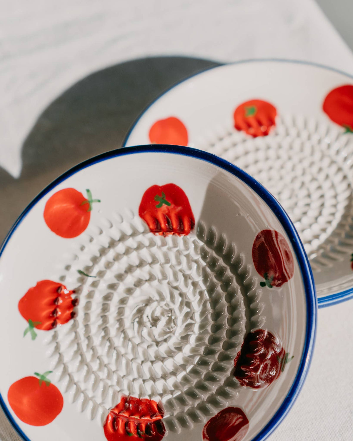 Zwei Teller der Serie „Alfar Tierra Cocida | Kleine Keramik Knoblauchreibe ‚Pomodoro‘“, handgefertigt in Spanien mit blauen Rändern und Tomatenmotiven sowie strukturierten spiralförmigen Mittelpartien, sind auf einer weißen Fläche ausgestellt, auf der das Sonnenlicht sanfte Schatten wirft.