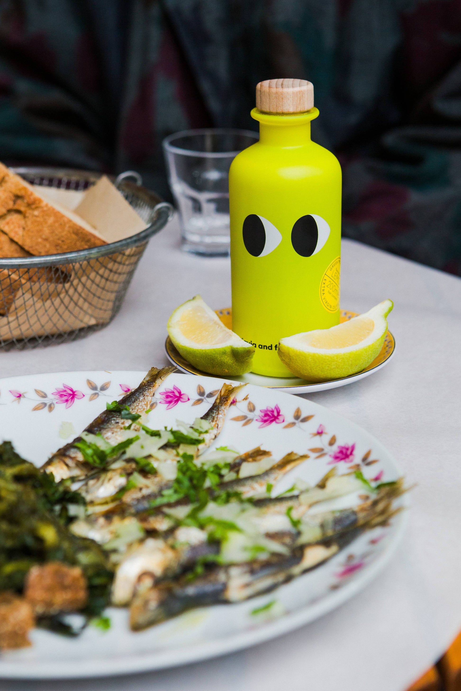 Ein Teller mit gegrillten Sardinen mit Kräutern steht neben Zitronenscheiben, einer leuchtend gelben Flasche "yiayia and friends | Natives Olivenöl extra mit Zitrone 200 ml" mit Augengrafik, einem Glas Wasser und einem Korb mit Brotscheiben.