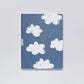Ein Notizbuch von nuuna | „Head In The Clouds“ Graphic L A5+ von nuuna mit handgezeichneten weißen Wolkenillustrationen auf einem blauen Einband aus recyceltem Denim und Premium-Papier, der auf einem schlichten weißen Hintergrund prangt.