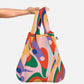Eine Hand hält die Notabag | 'Flow' Kombination aus Tasche & Rucksack von Notabag, die bunte abstrakte Wirbel in Pink, Rot, Grün, Orange, Lila und Gelb auf einem hellgrauen Hintergrund zeigt.