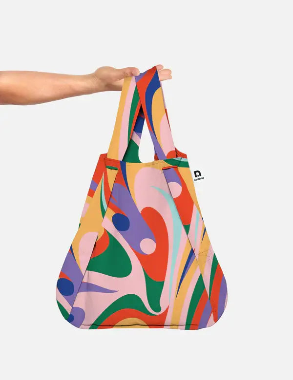Eine Hand hält die Notabag | 'Flow' Kombination aus Tasche & Rucksack von Notabag, die bunte abstrakte Wirbel in Pink, Rot, Grün, Orange, Lila und Gelb auf einem hellgrauen Hintergrund zeigt.