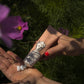 Eine Hand mit rotem Nagellack hält eine Tube der Archivist Gallery | 'Kirschblüte' Handcreme, die ein illustriertes Gesicht zeigt, während die andere die vegane Creme in einem Garten mit grünem Laub und rosa Blüten verteilt.