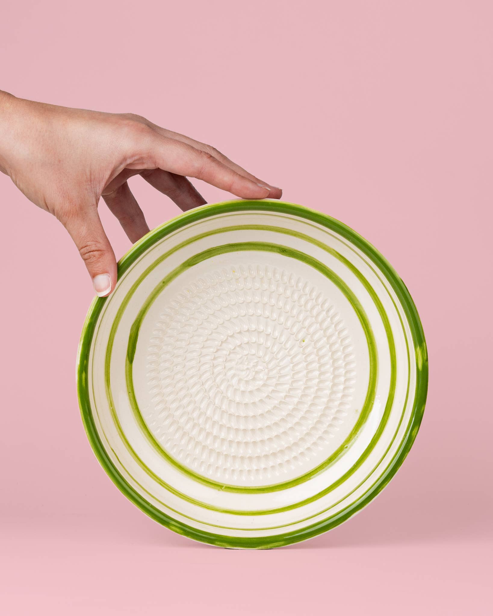 Eine Hand hält die „Alfar Tierra Cocida Große Keramik-Reibplatte ‚Olive‘“ – eine weiße Keramikplatte mit konzentrischen grünen Streifen und einer strukturierten Mitte, die als elegante mediterrane Küchenhilfe dient – vor einem rosa Hintergrund.