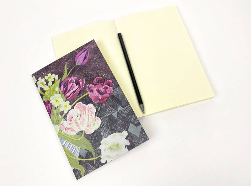 Ein aufgeschlagenes Angie Lewin | Notizbuch 'Tulpen und Narzissen' von Art Angels mit leeren Seiten und einem schwarzen Stift liegt neben einem geschlossenen floralen Notizbuch, alles auf weißem Hintergrund.