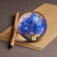 Der Hafod Grange | Briefbeschwerer mit Kornblume von Hafod Grange mit blauer Blüte liegt auf einem braunen Umschlag neben einem Holzbleistift-ein stilvolles Schreibtisch-Accessoire und ideales Geschenk für Gartenliebhaber.