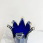 Die „Murano Dream Große Giusy“-Vase aus blauem, originalem Murano-Glas (H 32, Ø 20 cm) mit gewelltem Rand und transparenten Kanten steht auf weißem Marmor vor einem schlichten Hintergrund – ein Meisterwerk handgefertigter italienischer Glaskunst.