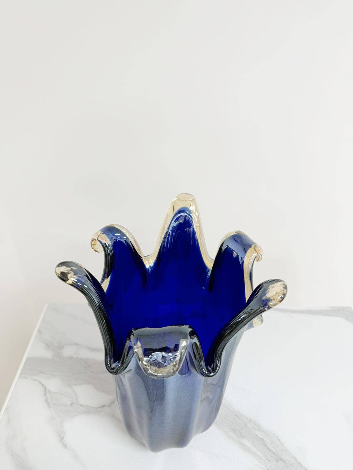 Die „Murano Dream Große Giusy“-Vase aus blauem, originalem Murano-Glas (H 32, Ø 20 cm) mit gewelltem Rand und transparenten Kanten steht auf weißem Marmor vor einem schlichten Hintergrund – ein Meisterwerk handgefertigter italienischer Glaskunst.