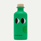 Eine kleine grüne 200-ml-Flasche "yiayia and friends | Natives Olivenöl extra mit Basilikum" mit zwei Cartoon-Augen, einem Holzdeckel, dem Markennamen am Boden und einem runden Stempel - das perfekte Feinkost Basilikumöl für Ihre Küche.