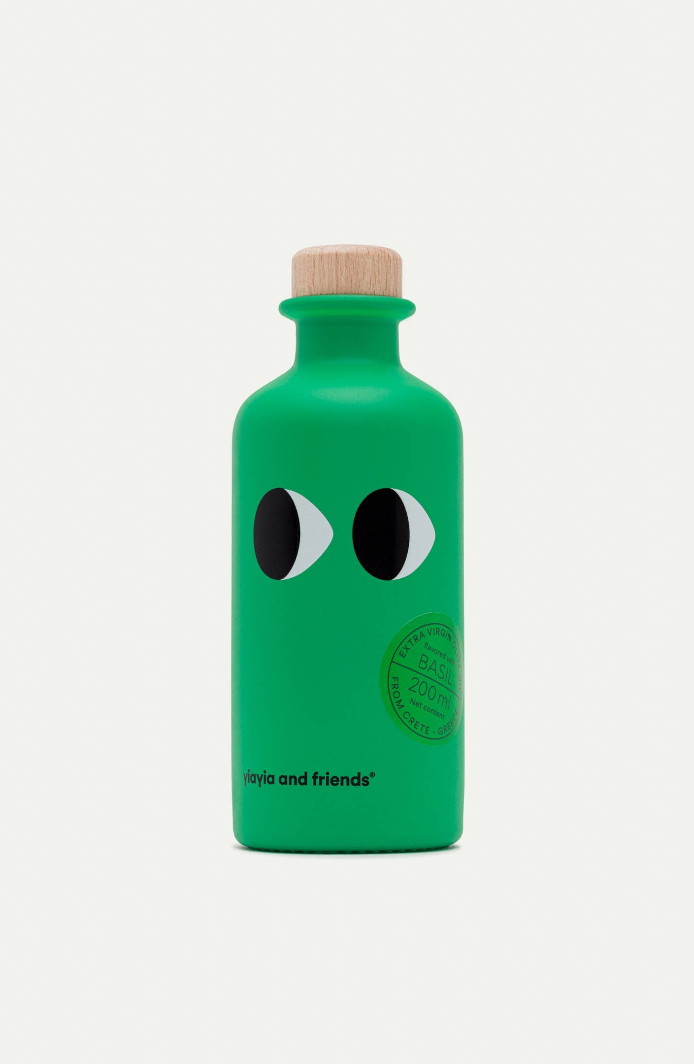 Eine kleine grüne 200-ml-Flasche "yiayia and friends | Natives Olivenöl extra mit Basilikum" mit zwei Cartoon-Augen, einem Holzdeckel, dem Markennamen am Boden und einem runden Stempel - das perfekte Feinkost Basilikumöl für Ihre Küche.