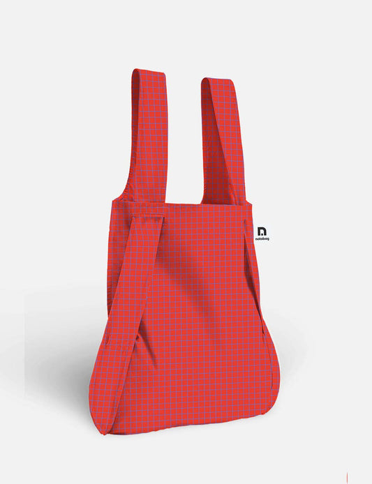 Notabag | Die wandelbare Tasche und der Rucksack „Beeren“ von Notabag ist eine rote Mehrwegtasche mit einem dezenten blauen Gittermuster, zwei breiten Tragegriffen und einem unauffälligen schwarz-weißen Seitenetikett, abgebildet vor einem schlichten hellen Hintergrund. Dieses innovative Design dient sowohl als Einkaufstasche als auch als Rucksack und bietet vielseitigen Stil und Komfort für den täglichen Gebrauch.