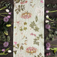 Der Koustrupco | Der Leinen-Tischläufer „Flora Danica Atlas“ im Format 145 x 45 cm von Koustrupco schmückt einen dunklen Holztisch, umgeben von rosa und weißen Tulpen, violettem Allium und üppigem Grün entlang der Ränder.