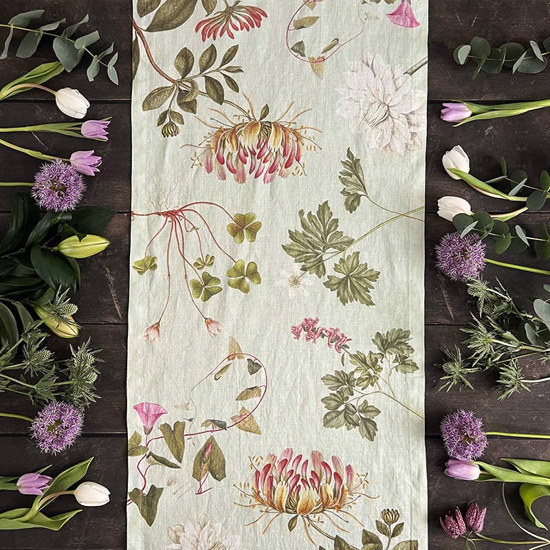 Der Koustrupco | Der Leinen-Tischläufer „Flora Danica Atlas“ im Format 145 x 45 cm von Koustrupco schmückt einen dunklen Holztisch, umgeben von rosa und weißen Tulpen, violettem Allium und üppigem Grün entlang der Ränder.