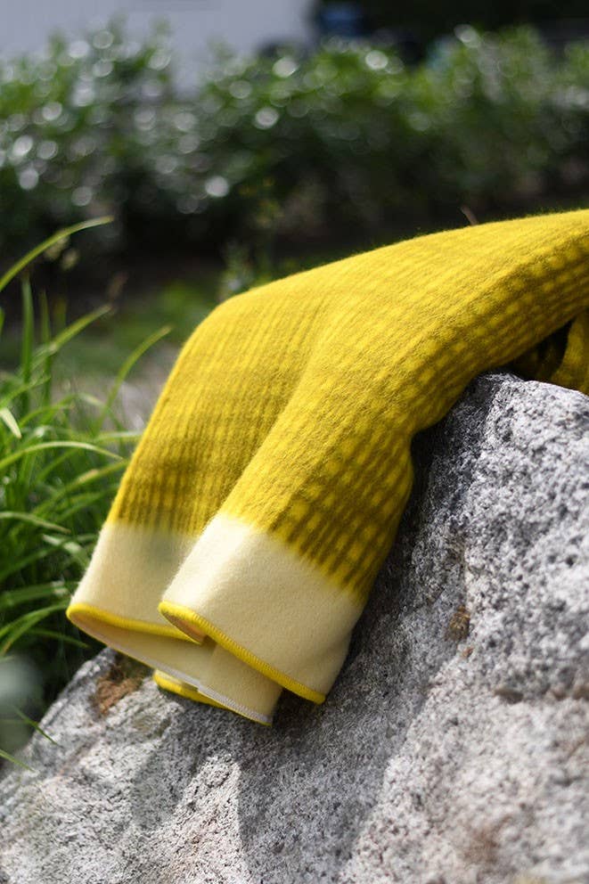 Die 'Darling' Gelbe Tagesdecke aus Lammwolle von Sofie Sjöström Design liegt auf rauem grauem Stein im Freien, umgeben von grünen Pflanzen und verschwommenem Laub im Hintergrund.