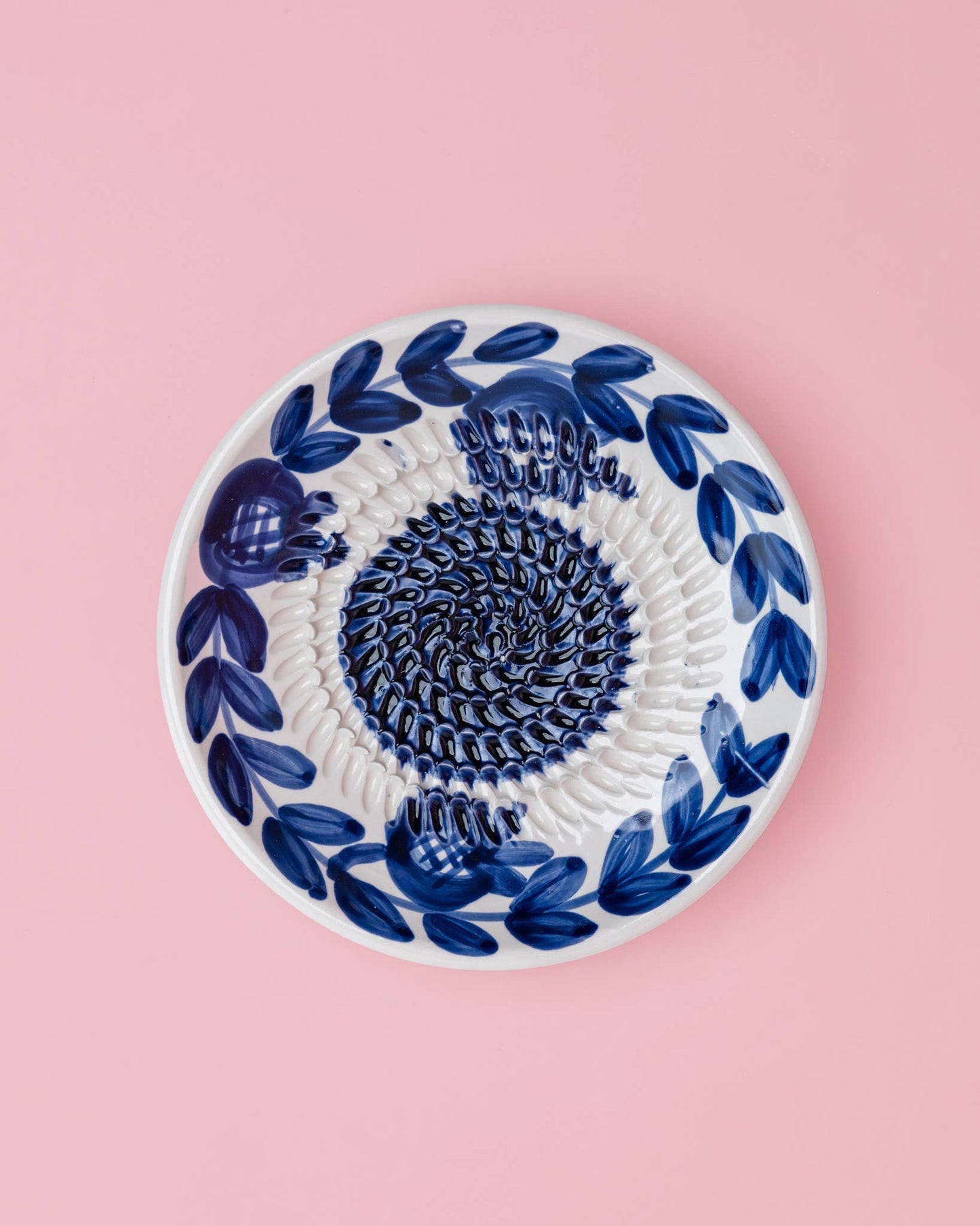Die Alfar Tierra Cocida | Kleine Keramik-Knoblauchreibeplatte „Nerja“ ist eine runde, weiße Keramikplatte mit traditionellen, handgemalten dunkelblauen Blattmotiven am Rand und einem strukturierten Spiralmuster in der Mitte, das sich vor einem hellrosa Hintergrund abhebt.
