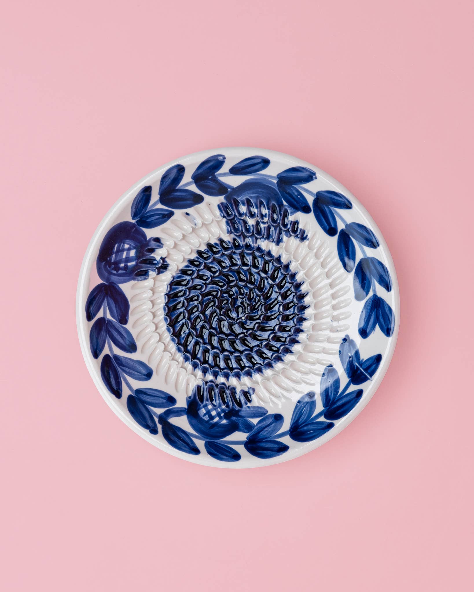 Die Alfar Tierra Cocida | Kleine Keramik-Knoblauchreibeplatte „Nerja“ ist eine runde, weiße Keramikplatte mit traditionellen, handgemalten dunkelblauen Blattmotiven am Rand und einem strukturierten Spiralmuster in der Mitte, das sich vor einem hellrosa Hintergrund abhebt.
