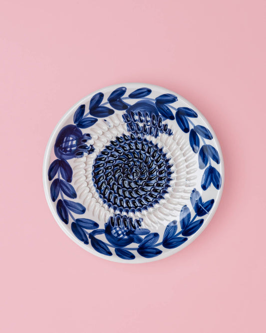 Die Alfar Tierra Cocida | Kleine Keramik-Knoblauchreibeplatte „Nerja“ ist eine runde, weiße Keramikplatte mit traditionellen, handgemalten dunkelblauen Blattmotiven am Rand und einem strukturierten Spiralmuster in der Mitte, das sich vor einem hellrosa Hintergrund abhebt.