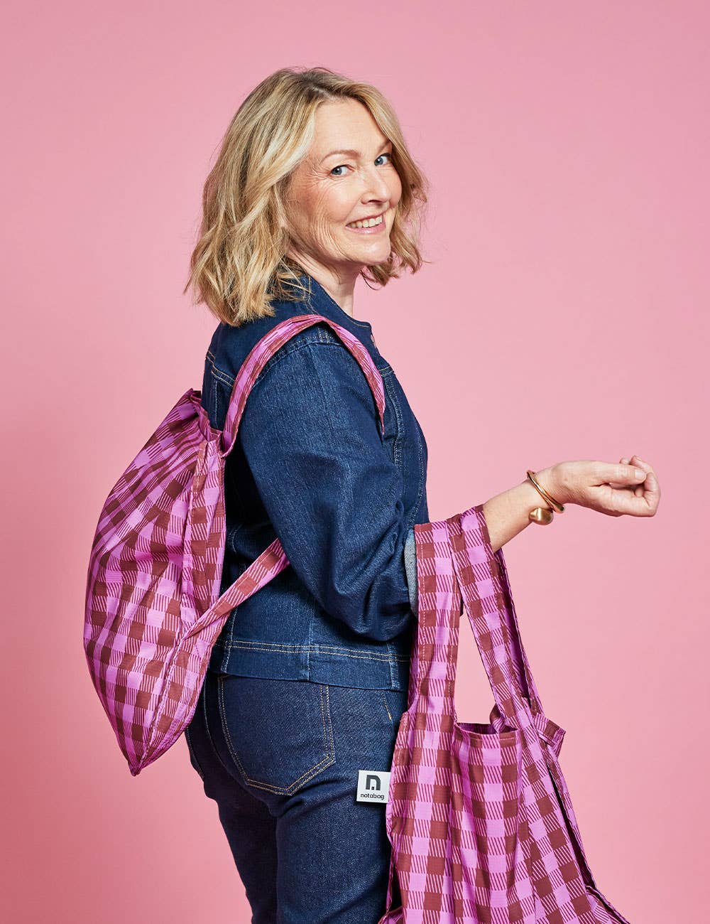Eine lächelnde Frau mit blonden Haaren, gekleidet in ein blaues Jeansoutfit, hält eine Notabag | Wandelbare Tasche und Rucksack „Kirsche“ von Notabag in der Hand – eine vielseitige Tasche im rosa Karomuster, die sich mühelos von einer Tragetasche in einen Rucksack verwandeln lässt –, während sie vor einem rosa Hintergrund steht.