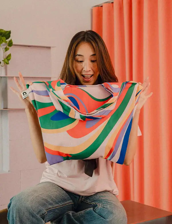 Eine Person mit langen braunen Haaren lächelt aufgeregt, während sie eine Notabag | Shopping Tote 'Flow' von Notabag, eine bunte wiederverwendbare Einkaufstasche, vor einem orangefarbenen Vorhang hält.